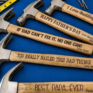 May include: Several claw hammers with wooden handles and metal heads are arranged on a blue surface. The handles are engraved with phrases like "Happy Father's Day" and "If Dad Can't Fix It, No One Can!"