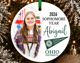 Könnte beinhalten: Ein rundes, weißes Keramikornament mit einem Foto einer Person mit langem braunem Haar in einem violetten Karohemd. Der Text lautet 2026 SOPHOMORE YEAR Abigail und OHIO UNIVERSITY, ergänzt durch eine grüne Wimpelgrafik mit dem Logo der Ohio University.