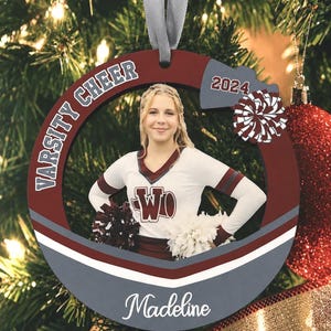 K&ouml;nnte beinhalten: Ein runder, kastanienbrauner und grauer Holzanh&auml;nger mit einem Foto einer Cheerleaderin in wei&szlig;er Uniform, beschriftet mit Varsity Cheer, 2024 und dem Namen Madeline. Der Anh&auml;nger hat ein graues Band zum Aufh&auml;ngen und ein Pompon-Motiv.