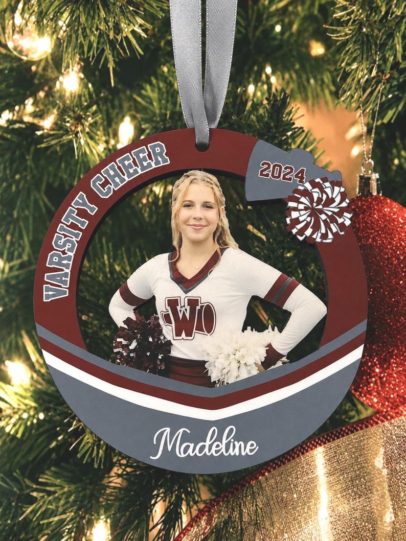 K&ouml;nnte beinhalten: Ein runder, kastanienbrauner und grauer Holzanh&auml;nger mit einem Foto einer Cheerleaderin in wei&szlig;er Uniform, beschriftet mit Varsity Cheer, 2024 und dem Namen Madeline. Der Anh&auml;nger hat ein graues Band zum Aufh&auml;ngen und ein Pompon-Motiv.