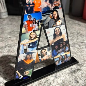 Puede incluir: Una letra A de acr&iacute;lico negro con un soporte, decorada con fotos de personas. Las fotos muestran personas en diferentes entornos, incluyendo un partido de f&uacute;tbol, una ceremonia de graduaci&oacute;n y un evento escolar. La letra es un regalo personalizado para alguien llamado A.