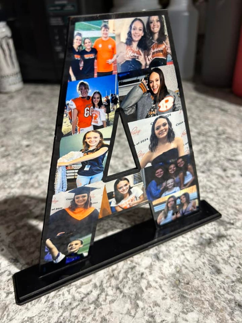 Puede incluir: Una letra A de acr&iacute;lico negro con un soporte, decorada con fotos de personas. Las fotos muestran personas en diferentes entornos, incluyendo un partido de f&uacute;tbol, una ceremonia de graduaci&oacute;n y un evento escolar. La letra es un regalo personalizado para alguien llamado A.