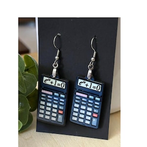 May include: A pair of silver earrings featuring a calculator design with the mathematical equation "e^ix + 1 = 0" displayed on the screen. The earrings are made of metal and have a black and blue color scheme.
