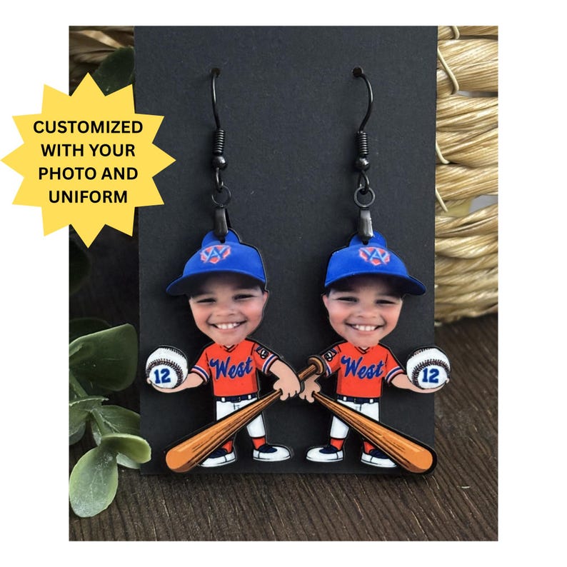 May include: A pair of dangle earrings featuring a cartoon baseball player with a smiling face, wearing a blue cap and an orange jersey with the word West, holding a baseball with the number 12 and a wooden bat, with black metal hooks.