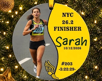 May include: A round ornament featuring a photograph of a runner on the left and a yellow background on the right. Text reads NYC 26.2 FINISHER, Sarah, 10/12/2026, #203, and -3:22:29-. A gold winged foot graphic is at the bottom right.