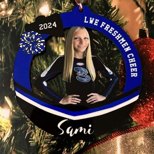 K&ouml;nnte beinhalten: Ein kreisf&ouml;rmiges blau-schwarzes Ornament mit einem Foto einer Cheerleaderin in schwarz-blauem Uniform-Oberteil mit der Nummer 3. Das Ornament zeigt den Text 2024, LWE FRESHMEN CHEER und den Namen Sami, akzentuiert durch ein blau-wei&szlig;es Pompon-Motiv.