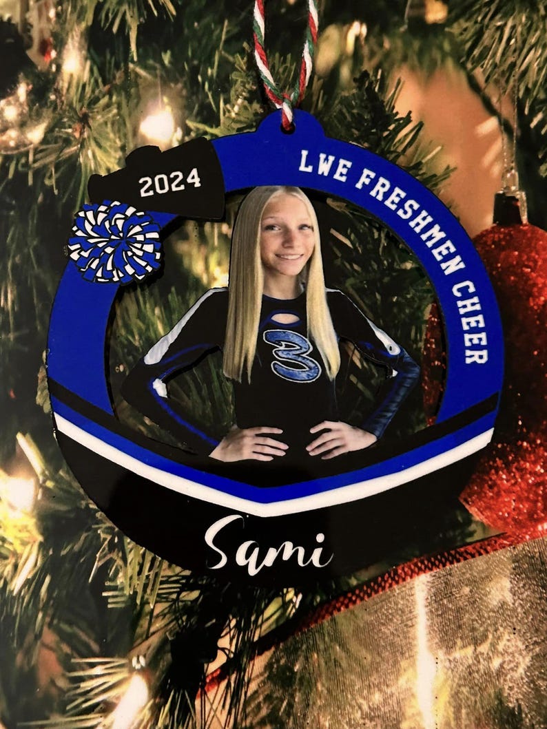 K&ouml;nnte beinhalten: Ein kreisf&ouml;rmiges blau-schwarzes Ornament mit einem Foto einer Cheerleaderin in schwarz-blauem Uniform-Oberteil mit der Nummer 3. Das Ornament zeigt den Text 2024, LWE FRESHMEN CHEER und den Namen Sami, akzentuiert durch ein blau-wei&szlig;es Pompon-Motiv.
