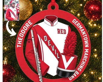 Könnte beinhalten: Ein runder roter Weihnachtsschmuck mit einer weiß-roten Marschkapellen-Uniformjacke mit dem Text RED DEVILS, einer silbernen Flöte und einem Hut mit roter Feder. Der Text THEODORE GERMANTOWN MARCHING BAND ist am Rand aufgedruckt.