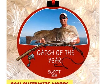 May include: A round red Christmas ornament featuring a photograph of a person holding a large fish. The ornament displays a graphic of a fishing rod and the text "CATCH OF THE YEAR", "SCOTT", and "2026" in white lettering against a red background.
