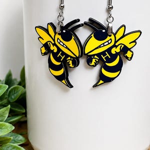 May include: A pair of dangle earrings featuring a yellow and black hornet mascot design with the letter H printed on the chest. The earrings are attached to silver-toned metal hooks and display a cartoon-style insect with wings and antennae.