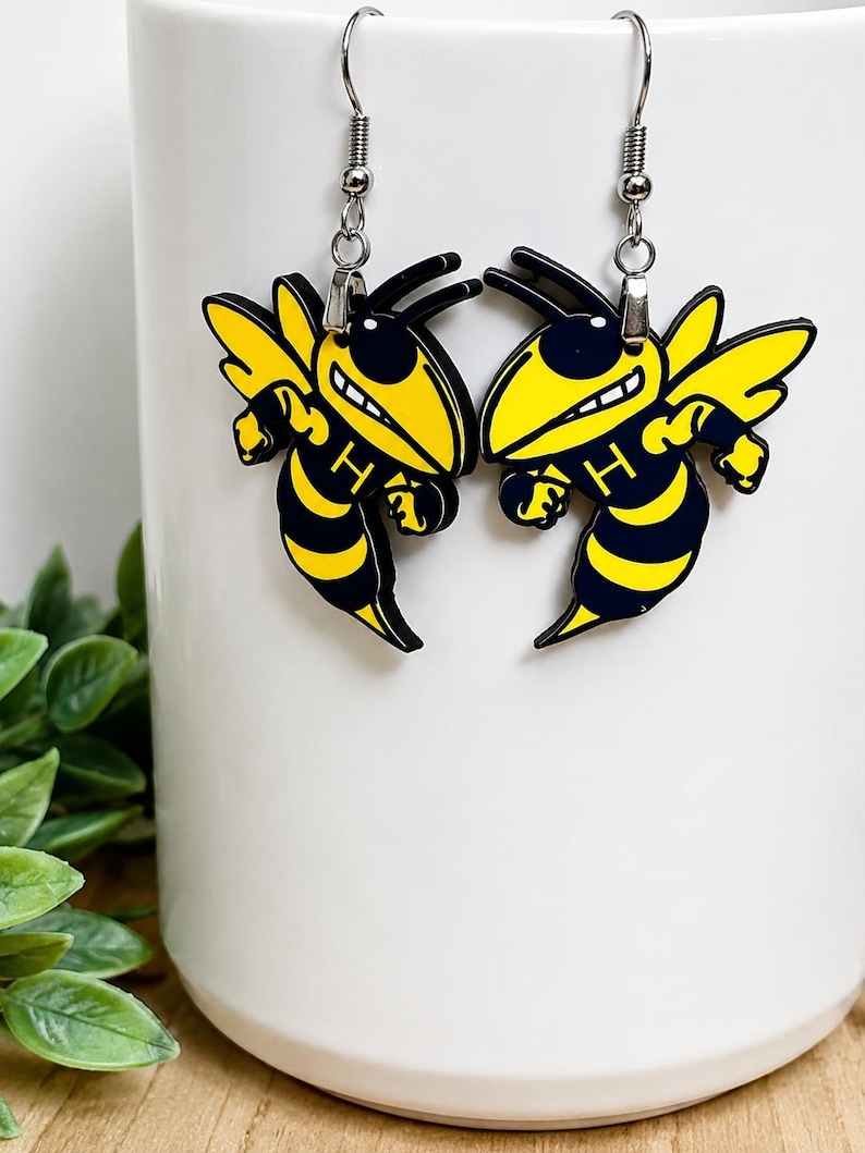 May include: A pair of dangle earrings featuring a yellow and black hornet mascot design with the letter H printed on the chest. The earrings are attached to silver-toned metal hooks and display a cartoon-style insect with wings and antennae.
