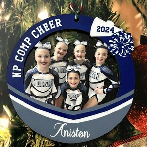 K&ouml;nnte beinhalten: Ein kreisf&ouml;rmiger blau-wei&szlig;er Weihnachtsschmuck mit einem Foto von f&uuml;nf Cheerleadern in wei&szlig;-dunkelblauen Uniformen. Der Text NP COMP CHEER, 2024 und der Name Aniston sind auf den Rahmen gedruckt, der mit einem wei&szlig;en Megafon und Pompon verziert ist.