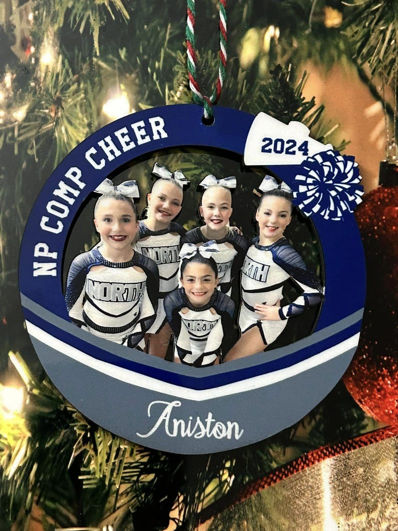 K&ouml;nnte beinhalten: Ein kreisf&ouml;rmiger blau-wei&szlig;er Weihnachtsschmuck mit einem Foto von f&uuml;nf Cheerleadern in wei&szlig;-dunkelblauen Uniformen. Der Text NP COMP CHEER, 2024 und der Name Aniston sind auf den Rahmen gedruckt, der mit einem wei&szlig;en Megafon und Pompon verziert ist.
