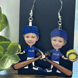 May include: A pair of dangle earrings featuring a cartoon baseball player character with a photographic face. The character wears a blue cap and a blue jersey with the name CALEB printed in white, holding a brown baseball bat and a yellow ball.