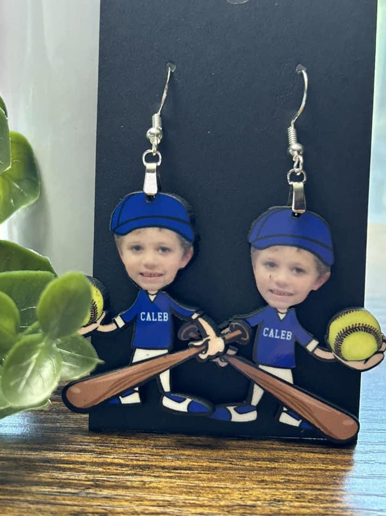 May include: A pair of dangle earrings featuring a cartoon baseball player character with a photographic face. The character wears a blue cap and a blue jersey with the name CALEB printed in white, holding a brown baseball bat and a yellow ball.