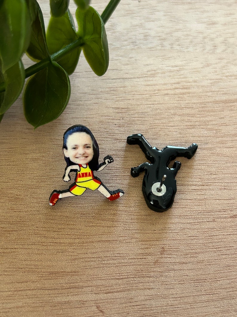 May include: A pair of stud earrings featuring a cartoon-style runner with a photographic face. The runner wears a yellow and red tank top with the name ANNA printed in red. The back of the earring shows a black silhouette with a metal post and clutch.