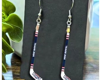 May include: A pair of dangle earrings shaped like miniature hockey sticks. The sticks are navy blue, white, and red with the text TRAVIS 2024 printed vertically on the shaft and the word REBELS printed near the blade, attached to silver hooks.