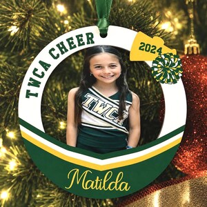 K&ouml;nnte beinhalten: Ein runder, wei&szlig;er Weihnachtsschmuck mit einem gr&uuml;nen Band zum Aufh&auml;ngen. Das Ornament zeigt ein Foto einer Cheerleaderin in gr&uuml;n-wei&szlig;er Uniform, den Text TWCA CHEER, das Jahr 2024, ein gelbes Megafon-Symbol, einen Pompon und den Namen Matilda.