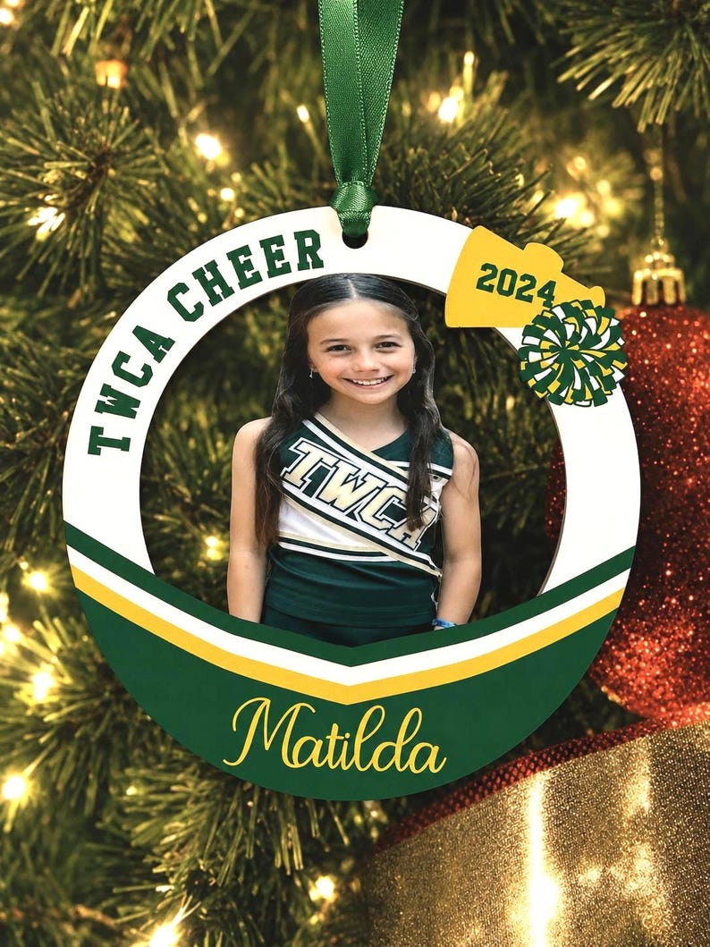 K&ouml;nnte beinhalten: Ein runder, wei&szlig;er Weihnachtsschmuck mit einem gr&uuml;nen Band zum Aufh&auml;ngen. Das Ornament zeigt ein Foto einer Cheerleaderin in gr&uuml;n-wei&szlig;er Uniform, den Text TWCA CHEER, das Jahr 2024, ein gelbes Megafon-Symbol, einen Pompon und den Namen Matilda.