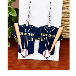 May include: A pair of dangle earrings featuring a navy blue baseball jersey design with the text BREW CREW and the number 10 in yellow. Each earring includes a light brown baseball bat and a white baseball with red stitching accents.