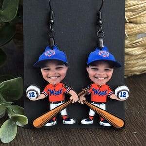 May include: A pair of dangle earrings featuring a cartoon baseball player with a smiling face, wearing a blue cap with a logo, an orange jersey with the word West, and white pants. The character holds a baseball with the number 12 and a bat.