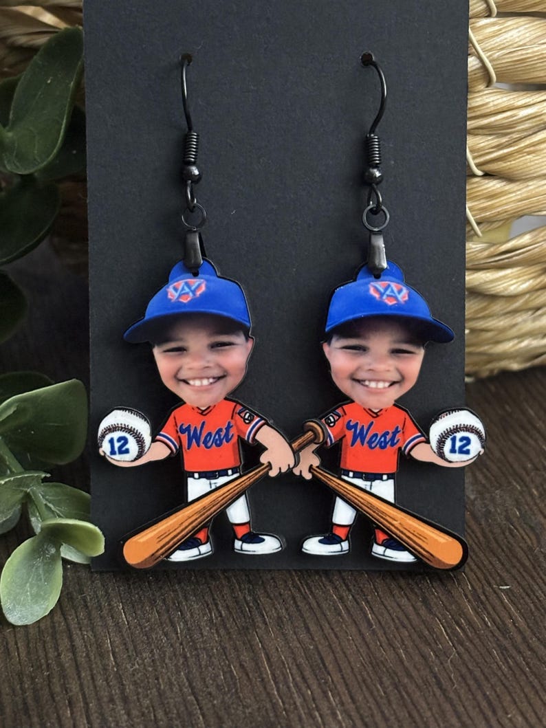 May include: A pair of dangle earrings featuring a cartoon baseball player with a smiling face, wearing a blue cap with a logo, an orange jersey with the word West, and white pants. The character holds a baseball with the number 12 and a bat.