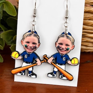 May include: A pair of dangle earrings featuring a cartoon-style softball player with a photographic face, wearing a blue jersey with the number 6, white pants, and a blue cap. The character holds a yellow softball and a wooden-colored bat.