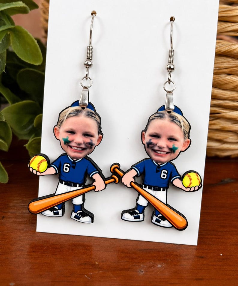 May include: A pair of dangle earrings featuring a cartoon-style softball player with a photographic face, wearing a blue jersey with the number 6, white pants, and a blue cap. The character holds a yellow softball and a wooden-colored bat.