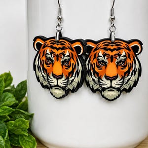 May include: A pair of dangle earrings featuring a detailed illustration of a tiger head. The tiger has vibrant orange fur with bold black stripes, white accents around the muzzle and ears, and piercing eyes. The earrings hang from silver-toned metal hooks.