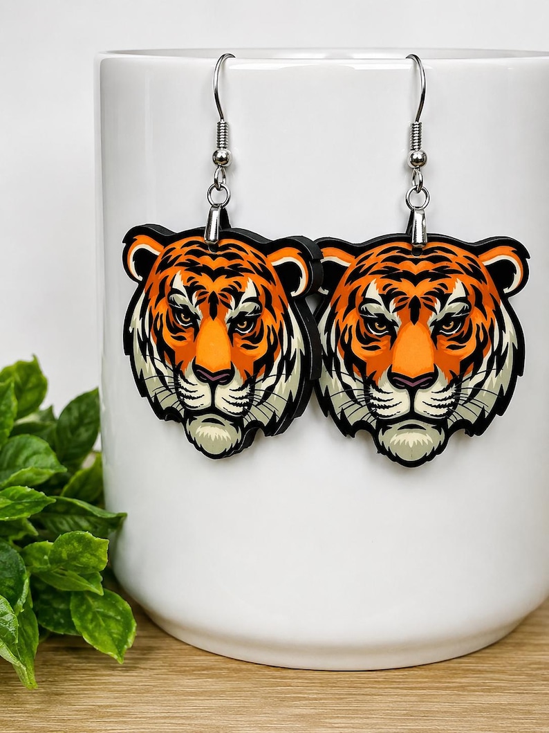 May include: A pair of dangle earrings featuring a detailed illustration of a tiger head. The tiger has vibrant orange fur with bold black stripes, white accents around the muzzle and ears, and piercing eyes. The earrings hang from silver-toned metal hooks.