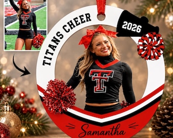 May include: A circular white and red Christmas ornament featuring a photo of a cheerleader in a black uniform with a red T logo. The ornament includes the text TITANS CHEER, the year 2026, a red bow, a red pom-pom, and the name Samantha in script.
