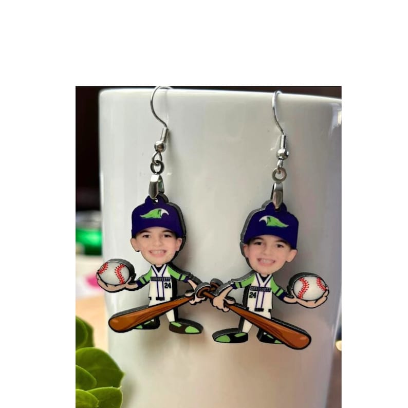 May include: A pair of dangle earrings featuring a cartoon-style baseball player with a smiling face, wearing a dark blue cap with a green logo, a white and green jersey with the number 24, holding a brown baseball bat and a white baseball.