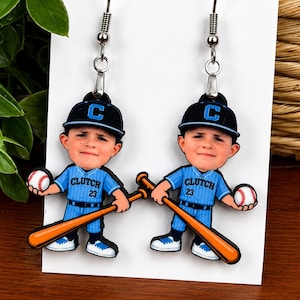 May include: A pair of dangle earrings featuring a cartoon baseball player wearing a blue uniform with the text CLUTCH 23, a black cap with a blue C, holding a baseball and a wooden bat. The earrings have silver-toned metal hooks for attachment.