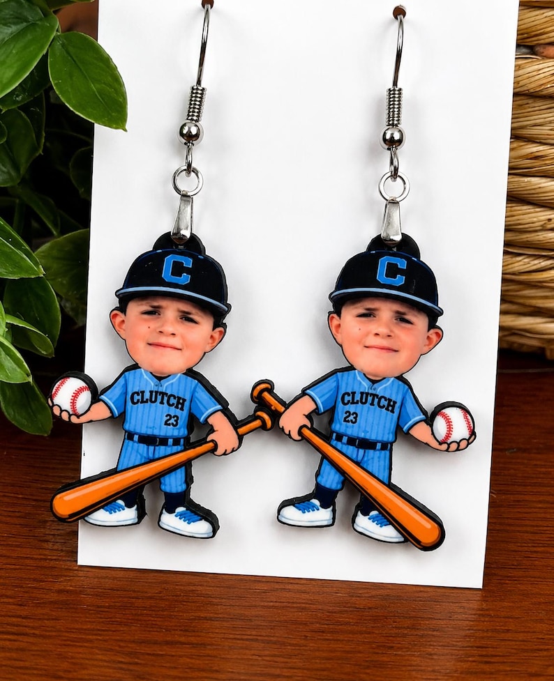 May include: A pair of dangle earrings featuring a cartoon baseball player wearing a blue uniform with the text CLUTCH 23, a black cap with a blue C, holding a baseball and a wooden bat. The earrings have silver-toned metal hooks for attachment.