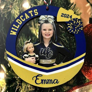K&ouml;nnte beinhalten: Ein kreisf&ouml;rmiger blau-goldener Weihnachtsschmuck mit einem Foto einer Cheerleaderin, die eine Puppe h&auml;lt. Der Rahmen zeigt den Text WILDCATS, 2024 und den Namen Emma in blauer Schrift. Ein kleiner blau-goldener Pompon ist oben rechts befestigt.