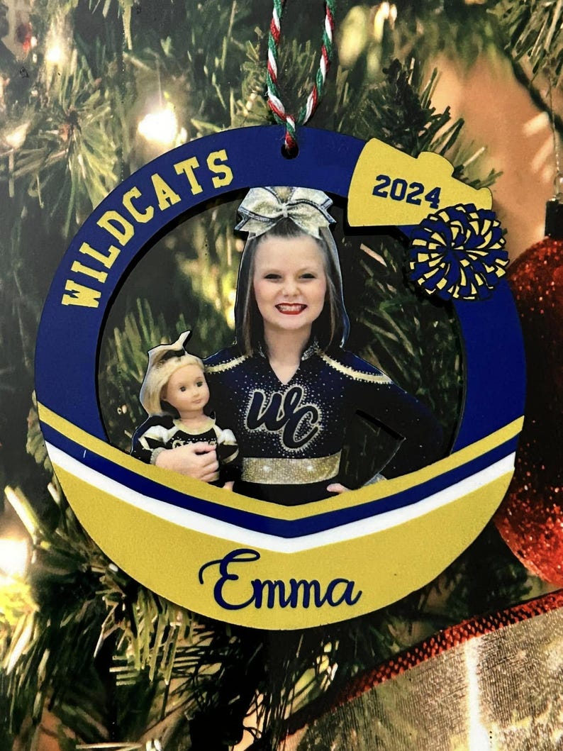 K&ouml;nnte beinhalten: Ein kreisf&ouml;rmiger blau-goldener Weihnachtsschmuck mit einem Foto einer Cheerleaderin, die eine Puppe h&auml;lt. Der Rahmen zeigt den Text WILDCATS, 2024 und den Namen Emma in blauer Schrift. Ein kleiner blau-goldener Pompon ist oben rechts befestigt.
