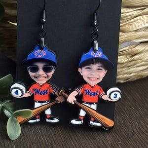 May include: A pair of dangle earrings featuring a cartoon baseball player character wearing an orange jersey with the word West, a blue cap, and white pants. Each figure holds a wooden baseball bat and a white baseball marked with the numbers 4 or 3.