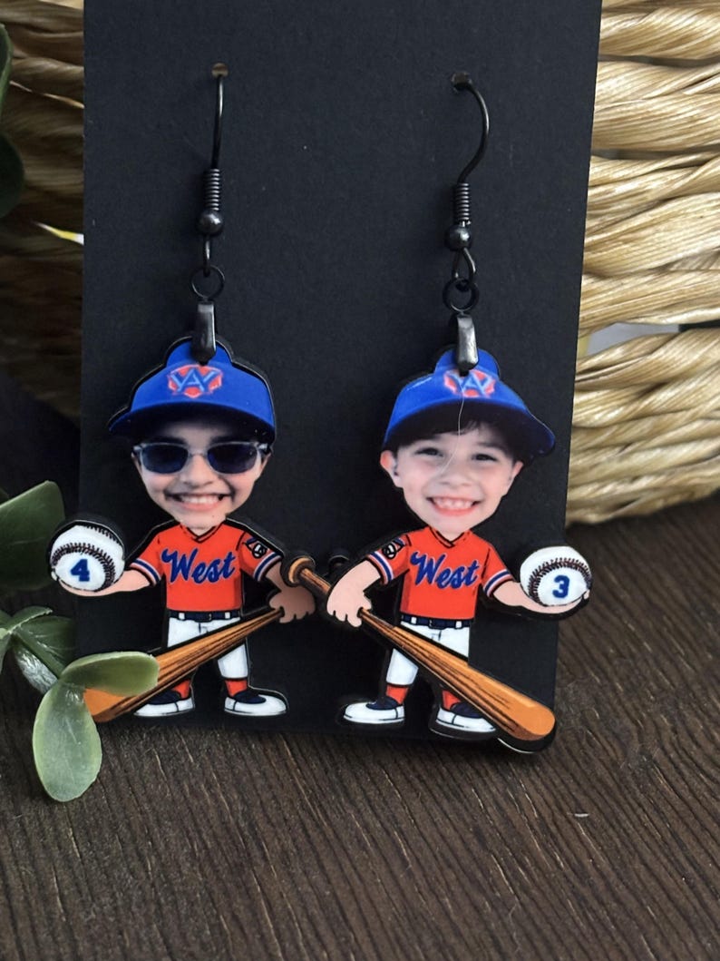 May include: A pair of dangle earrings featuring a cartoon baseball player character wearing an orange jersey with the word West, a blue cap, and white pants. Each figure holds a wooden baseball bat and a white baseball marked with the numbers 4 or 3.