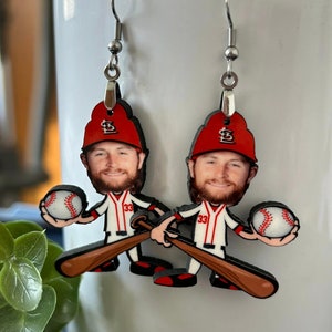 May include: A pair of dangle earrings featuring a cartoon-style baseball player with a beard, wearing a red cap with a white logo and a white jersey with the number 33 in red. The figure holds a white baseball and a brown wooden baseball bat.