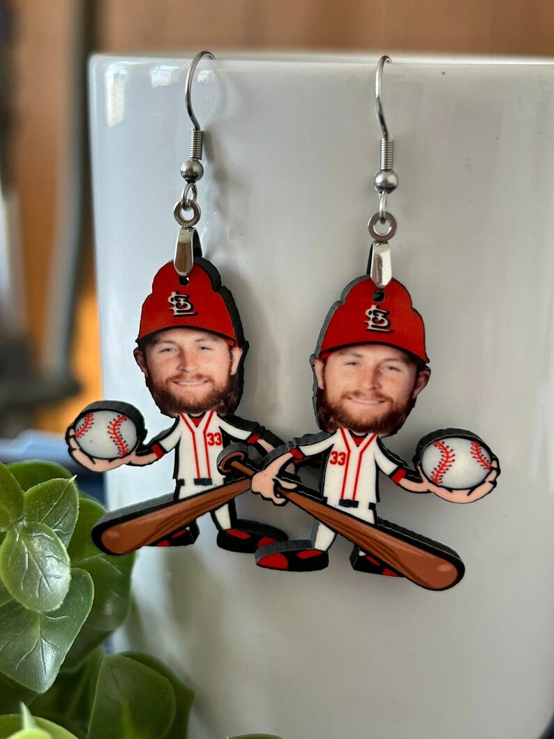 May include: A pair of dangle earrings featuring a cartoon-style baseball player with a beard, wearing a red cap with a white logo and a white jersey with the number 33 in red. The figure holds a white baseball and a brown wooden baseball bat.