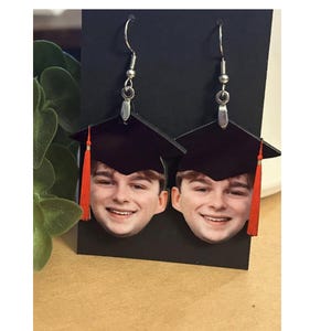 May include: A pair of graduation-themed earrings. Each earring features a smiling face beneath a black graduation cap with a red tassel. The earrings hang from silver-colored fish hook ear wires.