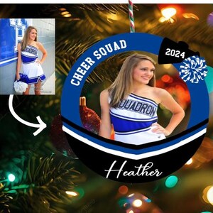 K&ouml;nnte beinhalten: Ein runder Weihnachtsschmuck mit blau-schwarzem Rand, der den Text CHEER SQUAD, 2024 und den Namen Heather tr&auml;gt. In der Mitte befindet sich ein Foto einer Person in einer blau-wei&szlig;en Cheerleader-Uniform, die einen blauen Pompon h&auml;lt.