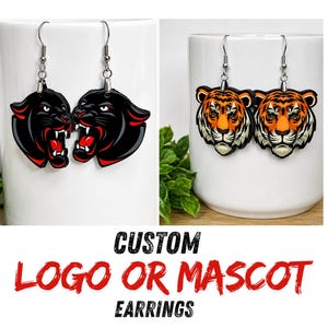 May include: Two pairs of dangling earrings featuring custom animal mascots. One pair displays black panther heads with bared teeth, while the other pair shows orange and black tiger heads. Each earring hangs from a silver-toned metal hook.