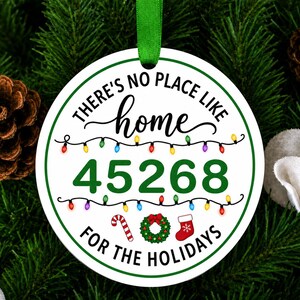 以下が含まれることがあります： 緑の縁取りと緑のリボンが付いた、丸い白いセラミック製のクリスマスオーナメント。テキストには「THERE'S NO PLACE LIKE home 45268 FOR THE HOLIDAYS」と書かれています。カラフルなイルミネーションライト、キャンディケイン、リース、赤い靴下が描かれています。