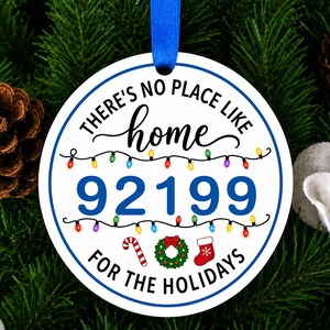 以下が含まれることがあります： 青い縁取りと青いリボンが付いた丸い白いクリスマスオーナメント。黒と青で「THERE'S NO PLACE LIKE home 92199 FOR THE HOLIDAYS」という文字が書かれており、カラフルなイルミネーション、キャンディケイン、リース、クリスマスストッキングのイラストが描かれています。