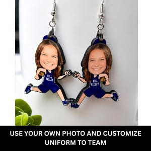 May include: A pair of dangling earrings featuring a custom photo of a person's face attached to a cartoon-style body wearing a dark blue athletic uniform with the text KILL VALLEY printed in white on the chest, suspended from silver-toned hooks.