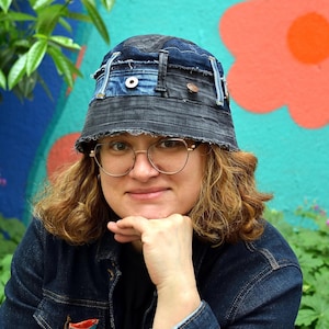 May include: A patchwork denim bucket hat, constructed from recycled jeans. The hat displays a mix of blue denim shades and visible stitching. The person in the image is wearing a denim jacket and glasses.