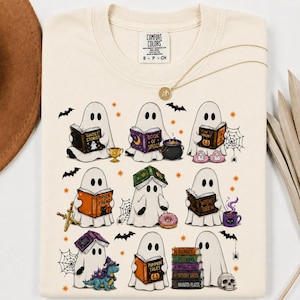 May include: A cream-colored t-shirt featuring a graphic print of nine white ghosts engaged in various activities, including reading books titled Ghost Stories, Book of Spells, and Pumpkin Tales, alongside illustrations of bats, spiders, and a skull.