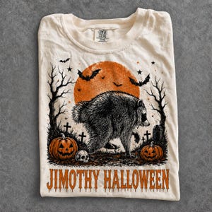 Pode incluir: Uma t-shirt de cor creme com um gráfico de um guaxinim a caminhar num cemitério com duas abóboras, um crânio, árvores despidas, morcegos e uma grande lua laranja. O texto JIMOTHY HALLOWEEN está impresso a laranja na parte inferior.