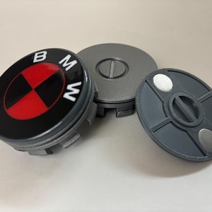 May include: Three gray and black BMW wheel center caps. One cap features the BMW logo in white on a black background with a red and black center. The other two caps are gray with a circular design.